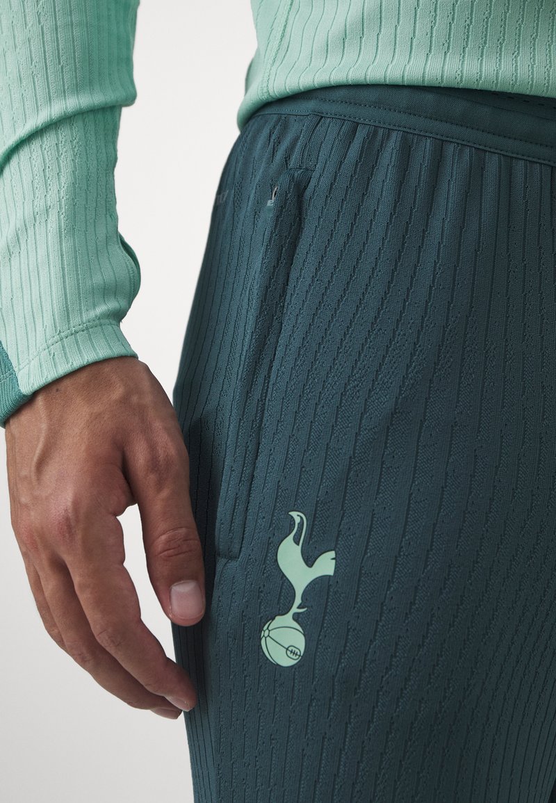 Un haut à manches longues en vert d'eau trouvé à côté d'un pantalon texturé en vert foncé. Les pantalons présentent un logo vert représentant un oiseau tenant un ballon de football.