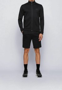 Giacca nera con zip e colletto alto, abbinata a pantaloni corti neri. Entrambi realizzati in tessuto liscio. Indossata con calze nere e scarpe sportive.