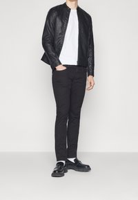 Giacca di pelle nera con colletto, t-shirt bianca, pantaloni neri slim-fit e scarpe nere con suola larga e hardware decorativo.