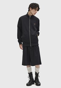 Felpa nera con zip e colletto alto, logo dorato e tasche anteriori, abbinata a pantaloni corti neri fino al ginocchio e stivali militari neri.