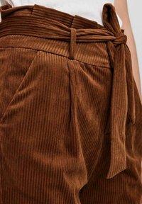Höga bruna kordfløjelsbyxor med veck, sidfickor, ett tygbälte knutet i en rosett och en vit topp instoppad.