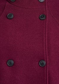 Burgundský vlněný kabát se zdvojeným zapínáním na černé knoflíky a hladkou texturou s jemnými stehy podél švů.