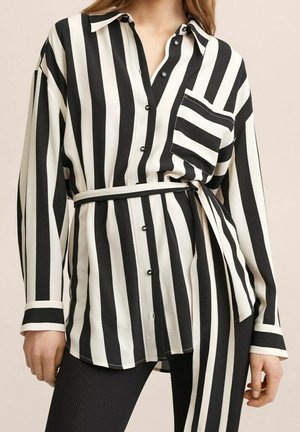 Femme portant une chemise à rayures verticales noires et blanches boutonnée, avec une ceinture assortie à la taille et une poche poitrine, associée à un pantalon noir côtelé.