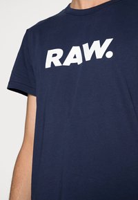 Marinblå bomulls-T-shirt med korta ärmar som har stor vit blocktext "RAW." på framsidan, slät textur och rundad halsringning.