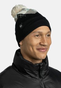 Gorro de punto negro con un pompón multicolor en la parte superior, que presenta patrones abstractos y una pequeña etiqueta con el logo. Llevado con una chaqueta negra.