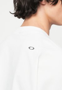 T-shirt blanc en coton, avec un col rond et un petit logo noir à l'arrière près de l'encolure. Texture lisse.