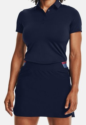 Kobieta ubrana w dopasowaną, granatową, krótkorękawową koszulkę polo i dopasowaną spódnicę z paskiem w paski, stojąca na białym tle.