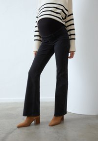 Gravid person i svarta graviditetsjeans, beige randig tröja och bruna klackiga ankelstövlar står på betonggolv.