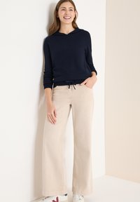 Granatowa bluza z kapturem z prążkowanymi mankietami, połączona z szerokimi spodniami w jasnobeżowym kolorze. Modelka nosi białe sneakersy z czerwonymi akcentami.