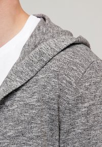 Primo piano di una persona che indossa una felpa con cappuccio grigia lavorata a maglia sopra una maglietta bianca a girocollo, su uno sfondo semplice.