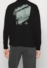 Felpa nera con maniche lunghe, caratterizzata da una grafica testurizzata color menta e branding Calvin Klein sul retro. Tessuto morbido, vestibilità rilassata.