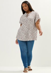 Kobieta stoi w białej bluzce w kwiaty z krótkim rękawem, niebieskich obcisłych dżinsach i beżowych sandałach na tle jednolitego jasnego tła.