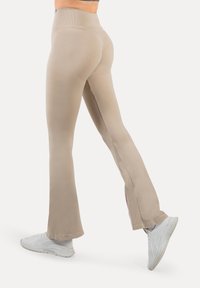 Beige flared legging met een gladde textuur en een hoge tailleband, gecombineerd met witte sportschoenen. Zijkantnaad zichtbaar.