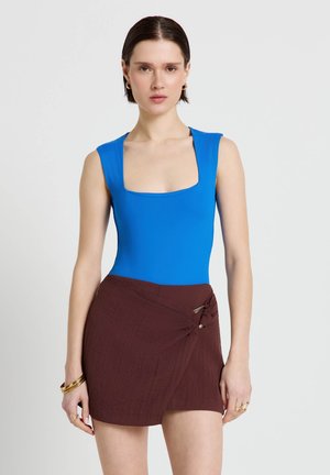 Femme portant un haut sans manches bleu vif avec un décolleté carré et une jupe portefeuille marron nouée sur le côté.