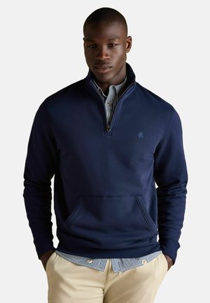 Marineblauer Pullover mit einem halb offenen Kragen, vorderer Kängurutasche und gerippten Bündchen. Getragen über einem gestreiften Hemd mit Kragen. Gleiche, glatte Textur.