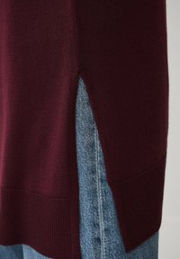 Bordeaux gebreide trui met zijsplitten, geribbelde zoom en een zachte textuur, gecombineerd met blauwe denim in een casual gelaagde stijl.