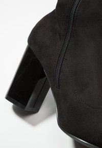 Bottine en daim noir avec un talon bloc brillant, équipée d'une fermeture éclair latérale et de détails de couture lisse. La forme est épurée et ajustée.