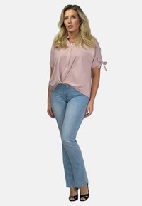 Różowy top z krótkim rękawem i skręconym przodem, zestawiony z jasnoniebieskimi spodniami dzwonami. Modelka ma na sobie czarne sandały na obcasie.