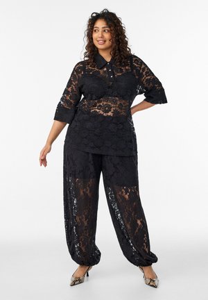 Žena oblečená v priesvitnom čiernom čipkovanom topánke a nohaviciach s golierom a gombíkmi, v kombinácii s hadím vzorom na vysokých podpätkoch, stojaca s rukou na boku.