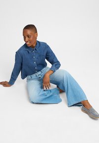 Dunkelblaue Jeanshemd mit Knöpfen und einer Brusttasche, kombiniert mit weiten hellblauen Jeans. Modell sitzt vor einem hellen Hintergrund.