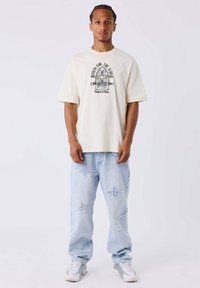 Crèmekleurig T-shirt met korte mouwen en grafische print; lichtblauwe jeans met sterrenborduursels; witte sneakers met grijze accenten.