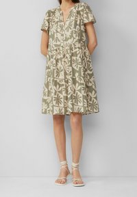 Eine Frau trägt ein knielanges, kurzärmliges Kleid mit grün-beige floralem Muster und weißen, schnürbaren flachen Sandalen.