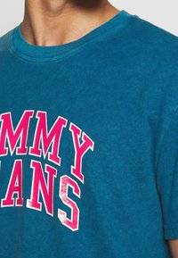 Camiseta de algodón en color azul verdoso con cuello redondo, que presenta un gran texto blanco y rosa "TOMMY JEANS" en el frente. Manga corta.