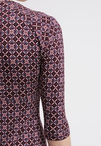 Långärmad klänning med ett geometriskt mönster i mörkröd, rosa och blått mot en mörk bakgrund; figurnära design med en mjuk textur.