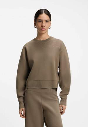 Mujer con cabello oscuro, con un suéter marrón de manga larga y pantalones a juego, de pie frente a un fondo blanco.