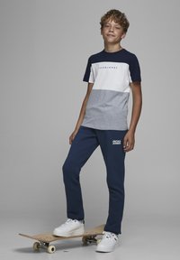 T-shirt a blocchi di colore blu e grigio con maniche corte e logo, abbinato a pantaloni sportivi blu navy con branding bianco, indossato con sneaker bianche.
