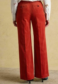 Plaša stila kordura bikses rūsas krāsā ar vertikālu ribotā raksta tekstūru. No aizmugures redzamas divas kabatas ar pogu aizdari.