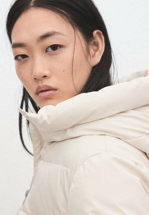 Pikowana, kremowa kurtka z wysokim kołnierzem, o gładkiej fakturze i widocznych szwach. Zbliżenie na kurtkę i twarz modela.