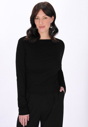 Kvinde med skulderlangt mørkt hår iført hængende øreringe, sort bluse med lange ærmer og sorte bukser, stående med den ene hånd i lommen.