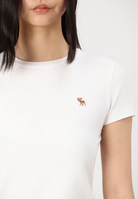 Camiseta blanca de algodón con mangas cortas que presenta un pequeño diseño de alce bordado en marrón en el pecho. Corte simple y ajustado.