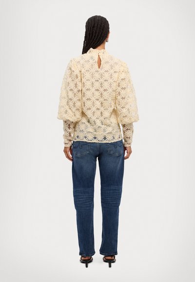 Lace-bluse i lys kremfarge med blomstermønster, puffede ermer og detaljer med nøkkelhull bak, kombinert med blå denimjeans.