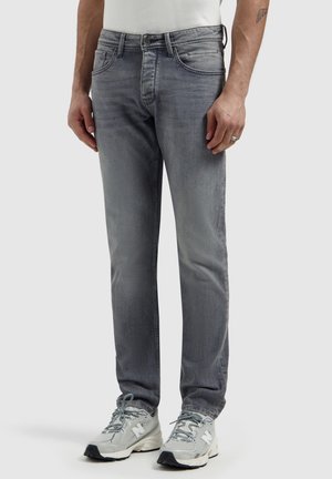 Mann trägt ausgewaschene graue Slim-Fit-Jeans und hellgraue New Balance Sneaker, steht vor einem schlichten hellen Hintergrund.