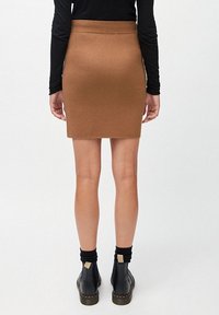 Vrouw in een zwart lang- mouwtop, een bruine rok tot op de knie, zwarte sokken en zwarte enkellaarsjes, staand tegen een effen achtergrond.