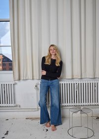 Top nero a maniche lunghe abbinato a jeans a gamba larga di colore azzurro chiaro. A piedi nudi su uno sfondo bianco, con tende e uno sgabello metallico.