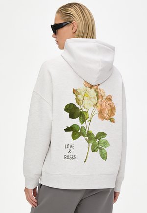 Persona che indossa occhiali da sole e una felpa chiara con grandi rose e la scritta "LOVE & ROSES" stampata sulla schiena, di spalle.