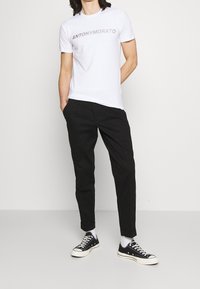 White t-shirt with "ANTONY MORATO" graphic, paired with black trousers and black sneakers. Model poses with one hand in pocket.