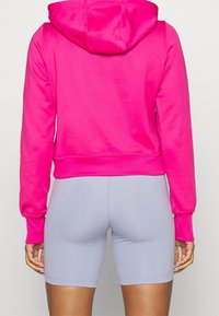 Personne portant un hoodie rose vif et un short ajusté gris clair, se tenant de dos contre un fond uni.