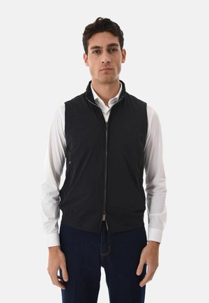 Uomo con indosso una camicia bianca a maniche lunghe e un gilet nero senza maniche con cerniera, in piedi contro uno sfondo chiaro e uniforme.