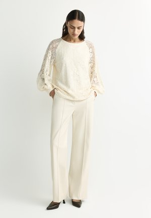 Woman with long dark hair wearing an off-white lace blouse with puffed sleeves, off-white wide-leg pants, and black pointed heels.