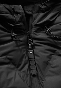 Giacca a vento nera con superficie texturizzata, dotata di una zip prominente, cordini regolabili e dettagli del logo sul tirante della zip e sulle cinghie.