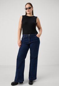 Top sans manches à côtes noires, jeans évasés bleu foncé avec poches frontales, porté avec des bottes à talons carrés noires et des lunettes de soleil foncées.