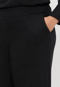 Czarne spodnie mają gładką teksturę z delikatnym połyskiem. Wyraźnym detalem jest kieszeń ozdobiona czarnymi koralikami wzdłuż krawędzi.