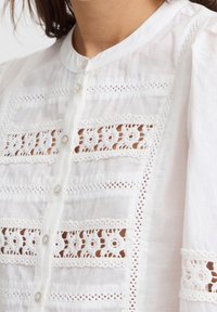 Blusa di cotone bianca con pannelli ricamati decorativi, colletto rotondo e chiusura a bottoni frontale. Accenti di pizzo testurizzato in tutto il capo.
