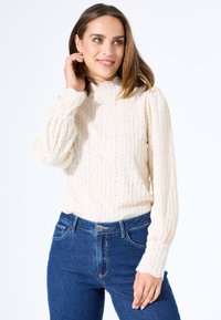 Crème-kleurige pullovertrui met een textuurpatroon, hoge hals en pofmouwen, gecombineerd met donkerblauwe spijkerbroek.