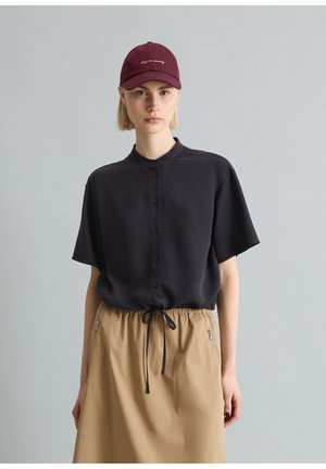 Młoda osoba nosząca bordową czapkę z różowym napisem, czarną koszulę na krótkie rękawy z guzikami oraz beżową spódnicę z kieszeniami na zamek z przodu.