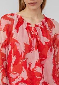 Blouse à motifs rouges et roses en tissu doux et léger, avec un col froncé et une bordure à volants. Se noue à l'avant.